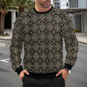A man wearing a black crewneck sweatshirt with a beige geometric circular pattern, standing outdoors on a street with palm trees and buildings in the background.