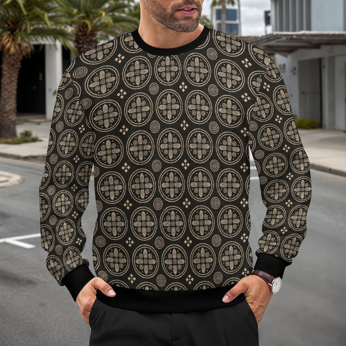 A man wearing a black crewneck sweatshirt with a beige geometric circular pattern, standing outdoors on a street with palm trees and buildings in the background.