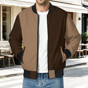 A man wearing a color-blocked bomber jacket with brown and tan panels, black ribbed collar, cuffs, and hem, layered over a white t-shirt and blue jeans, standing on a street in front of a cafe.