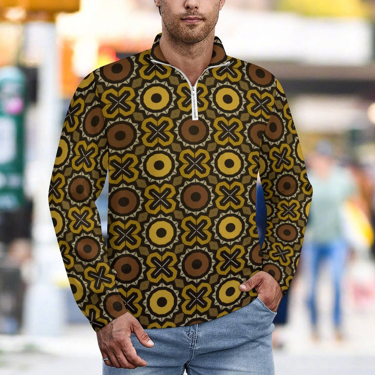 A man wearing a long-sleeve quarter-zip pullover with a bold retro geometric pattern in olive green, brown, and mustard yellow, paired with light blue jeans, set against a blurred outdoor street background.