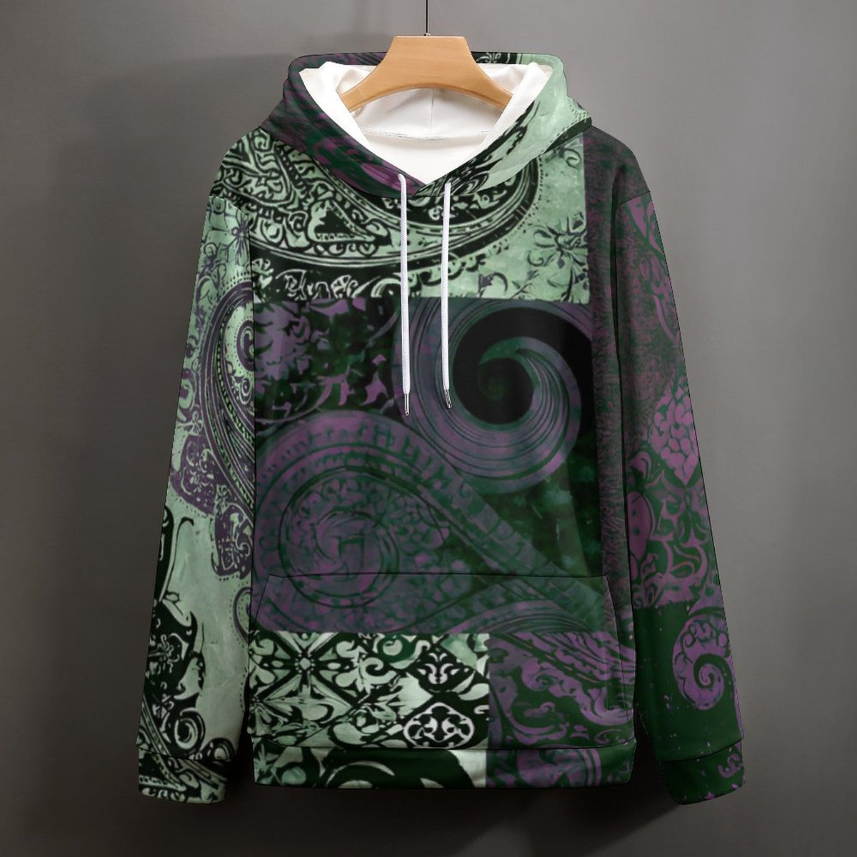 A pullover hoodie with a complex paisley and floral pattern in shades of green, purple, and black, hanging on a wooden hanger against a dark gray wall.