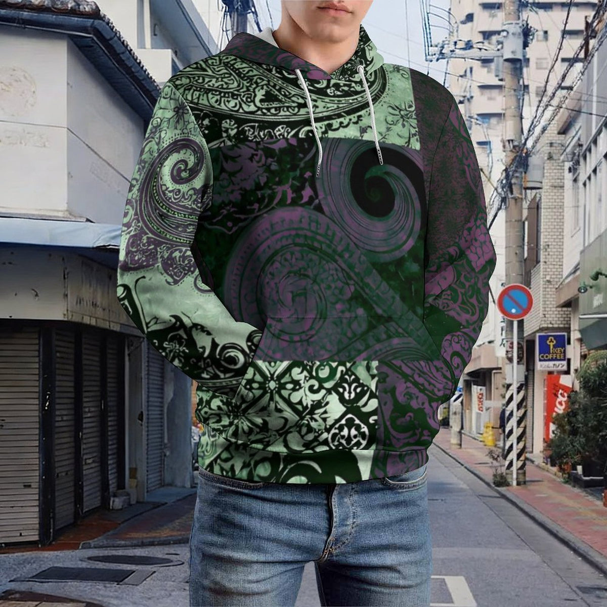 A person wearing a pullover hoodie with an all-over abstract paisley and swirl pattern in black, green, and purple, standing on a city street.