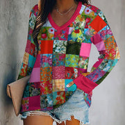 A woman wearing a long-sleeve V-neck patchwork blouse with a vibrant multicolor floral and geometric print, paired with denim shorts and holding a beige clutch bag against a gray textured wall.