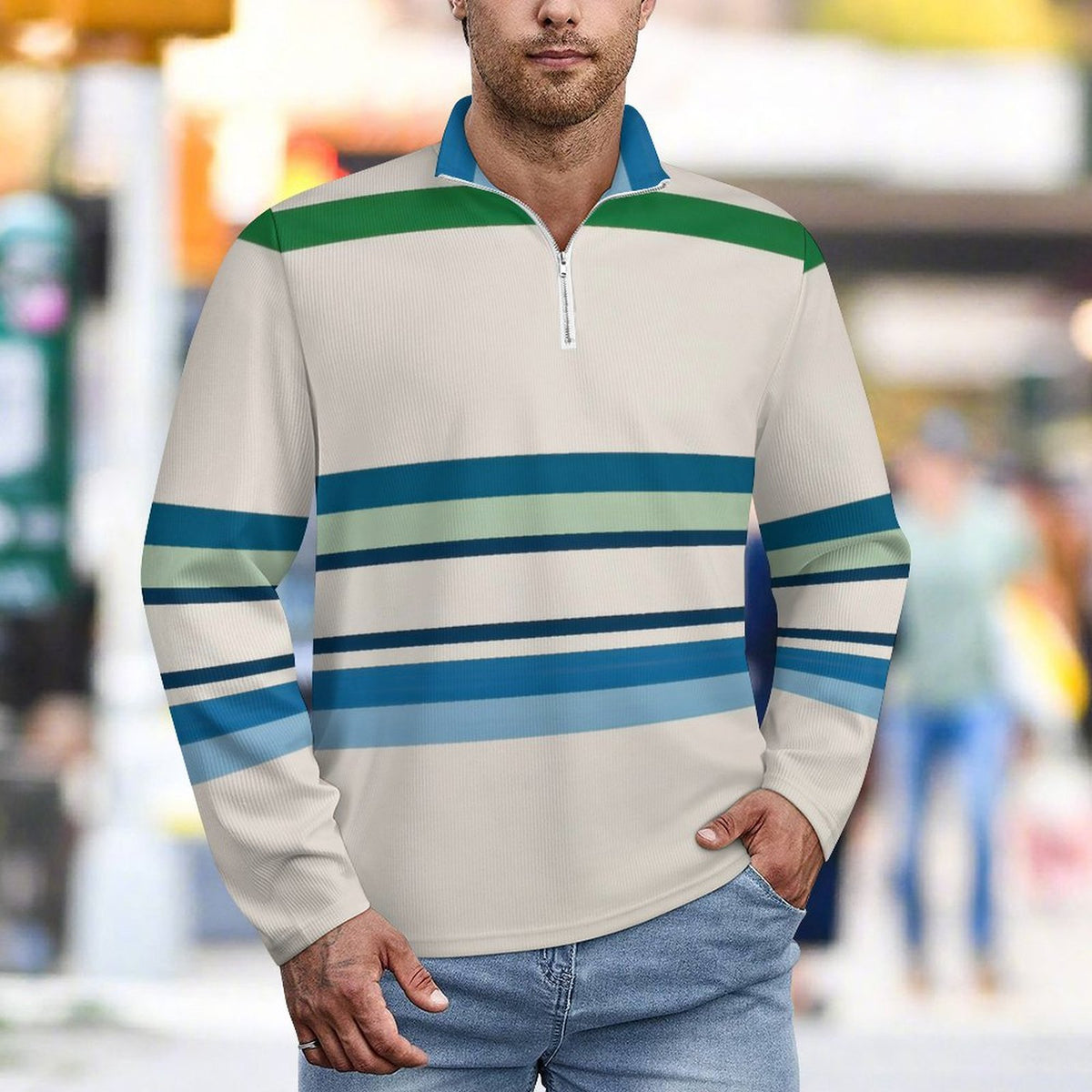 A man wearing a long-sleeve quarter-zip pullover with horizontal stripes in green, blue, and light teal on a cream base, paired with blue jeans, set against a blurred city street background.